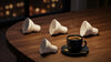 A steaming cup of coffee in a black cup sits on a wooden table with four scattered LED light bulbs. The scene is warm and cozy, set against blurred city lights.