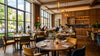 Modern restaurant interior with large windows, wooden floors, and elegant decor. Patrons dine at round tables, with soft lighting and a relaxed atmosphere.