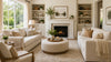A cozy, elegant living room with neutral tones. Features two sofas, a round ottoman, a fireplace, bookshelves, and large window. Soft natural light enhances warmth.